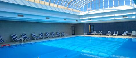 An indoor swimming pool with a glass ceiling, lounge chairs, and a tiled wall.
