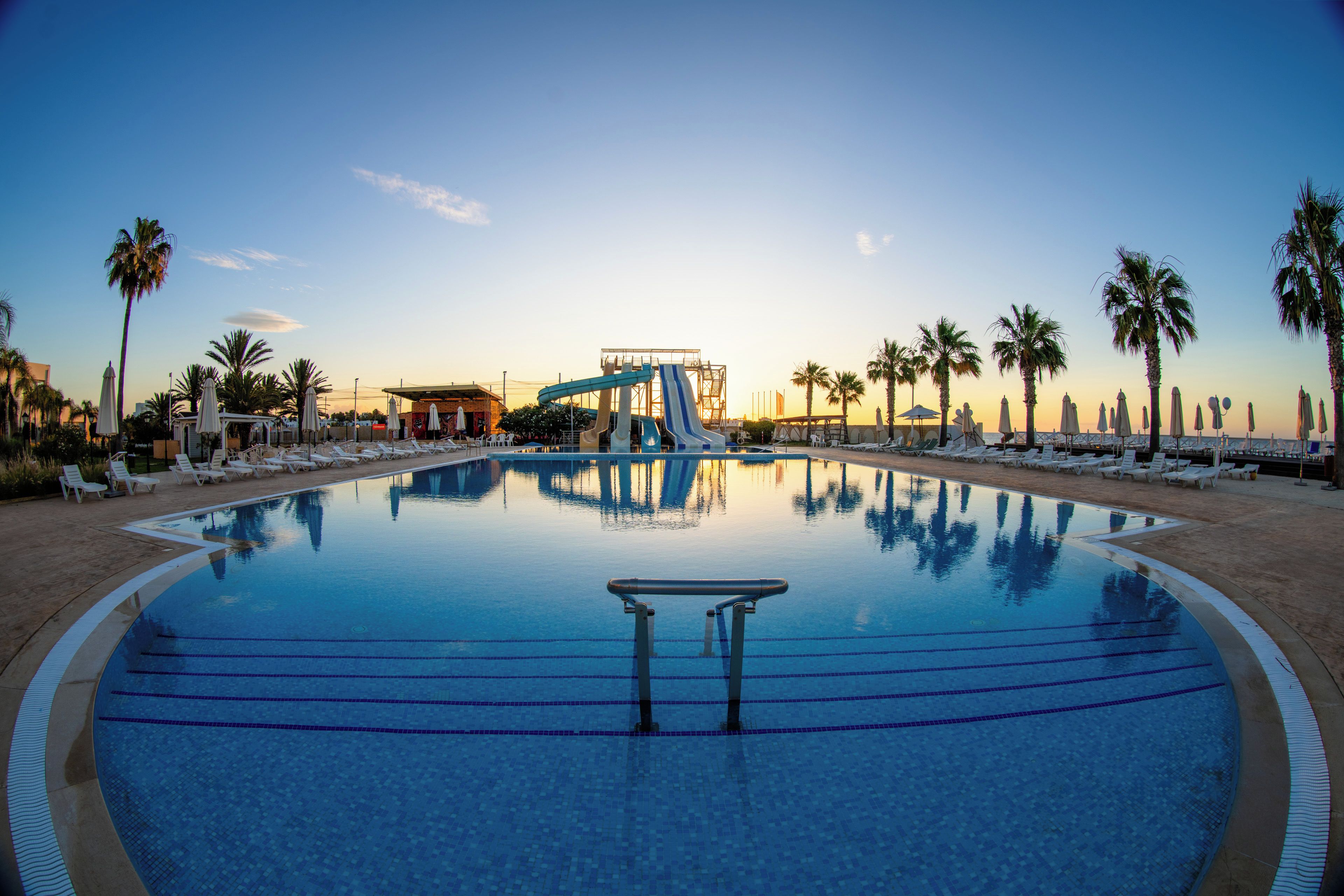 A swimming pool with a water slide, lounge chairs, and palm trees.