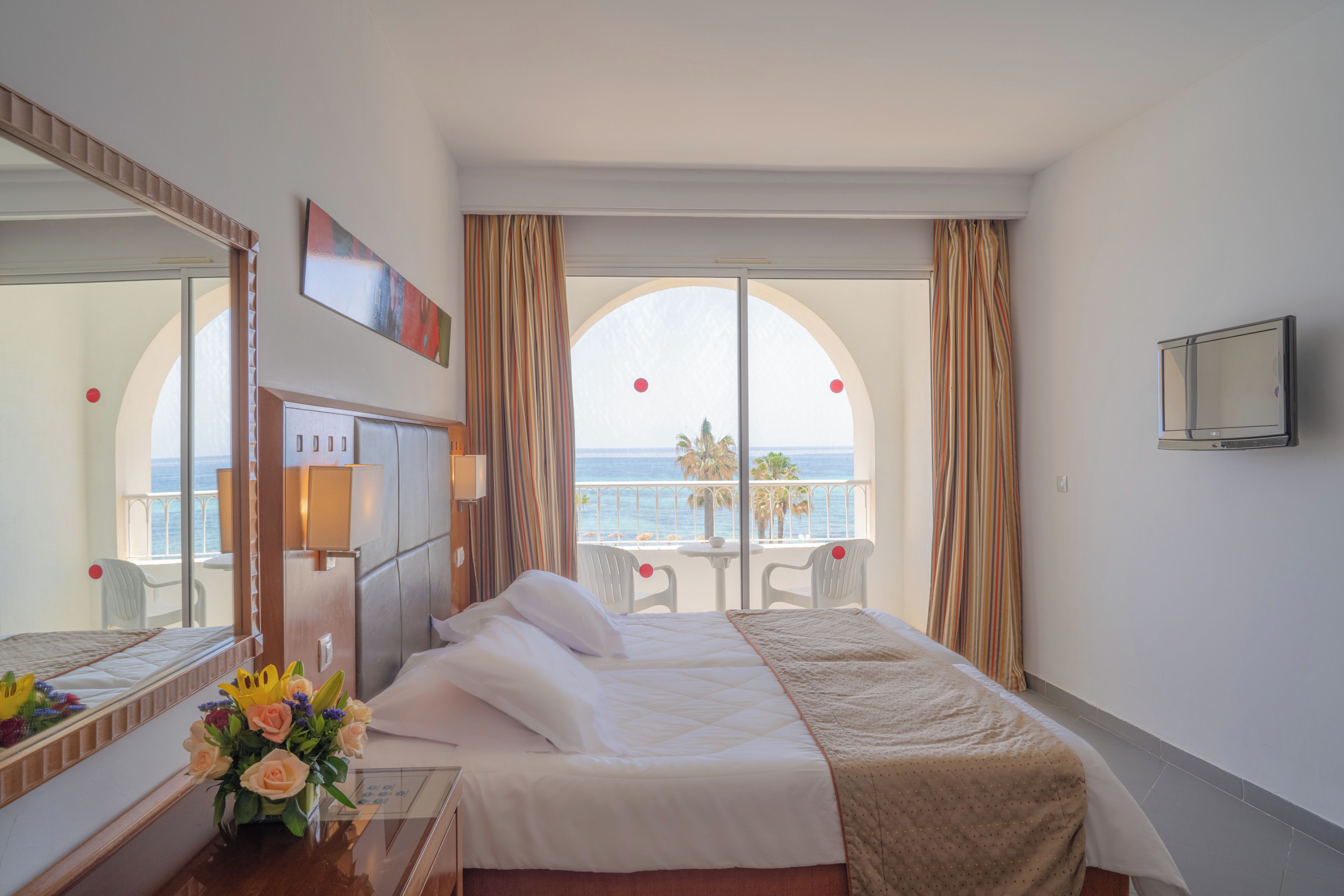 A hotel room with a bed, a desk, a mirror, a TV, and a view of the sea.