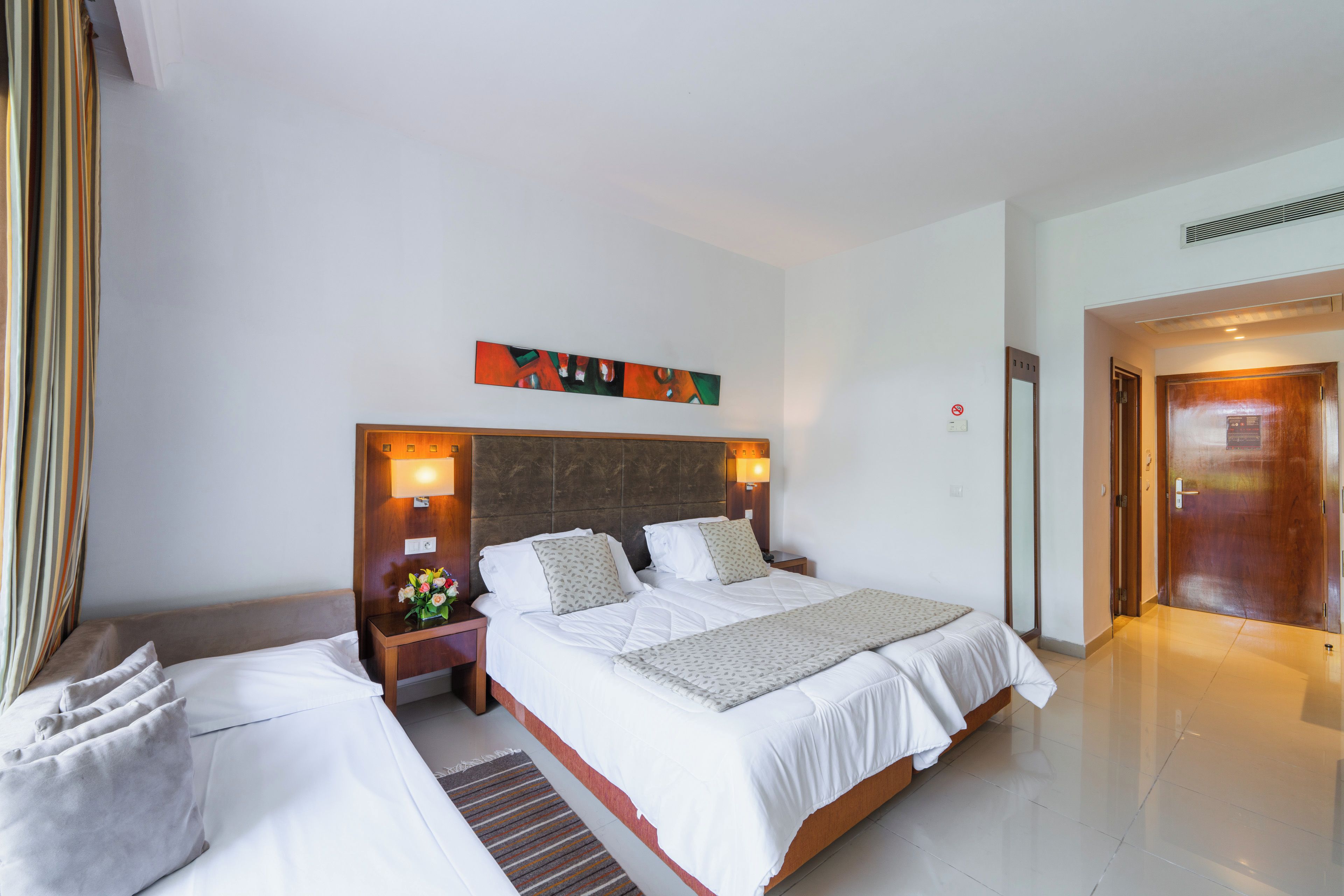 A hotel room with a large bed, a smaller bed, a nightstand with a vase of flowers, and a painting on the wall.