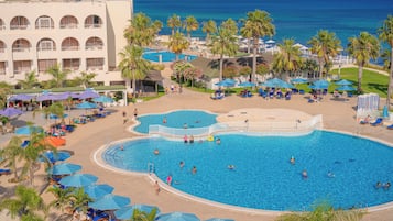 Una amplia piscina al aire libre rodeada de palmeras y sillas de descanso, con un edificio al fondo.