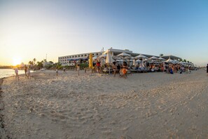 Une plage avec un grand bâtiment sur lequel on peut lire « COCO BEACH » en arrière-plan, de nombreux parasols et des gens qui profitent de la plage.