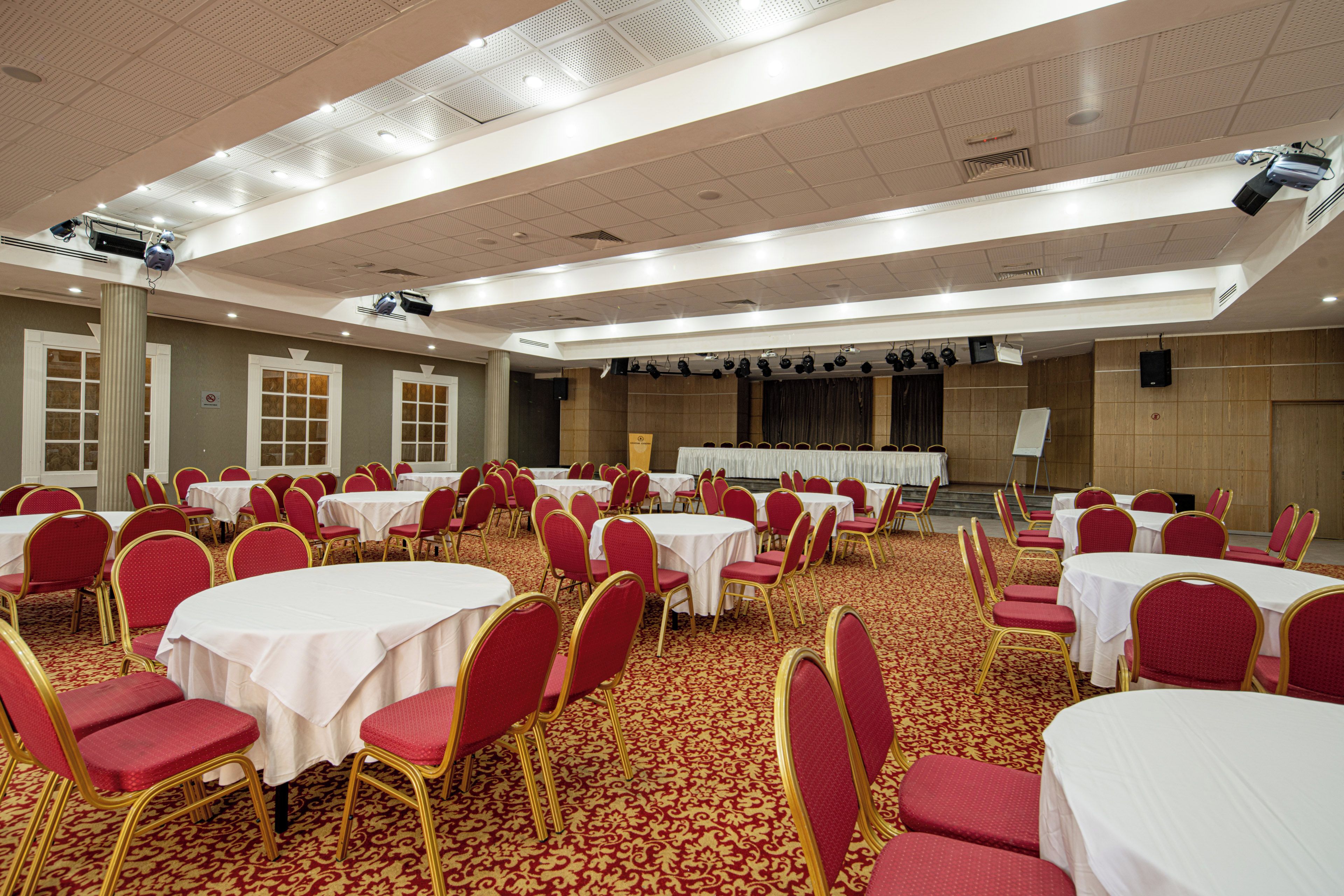 Un salón de banquetes con mesas redondas y sillas rojas, equipado con proyectores y luces instalados en el techo.