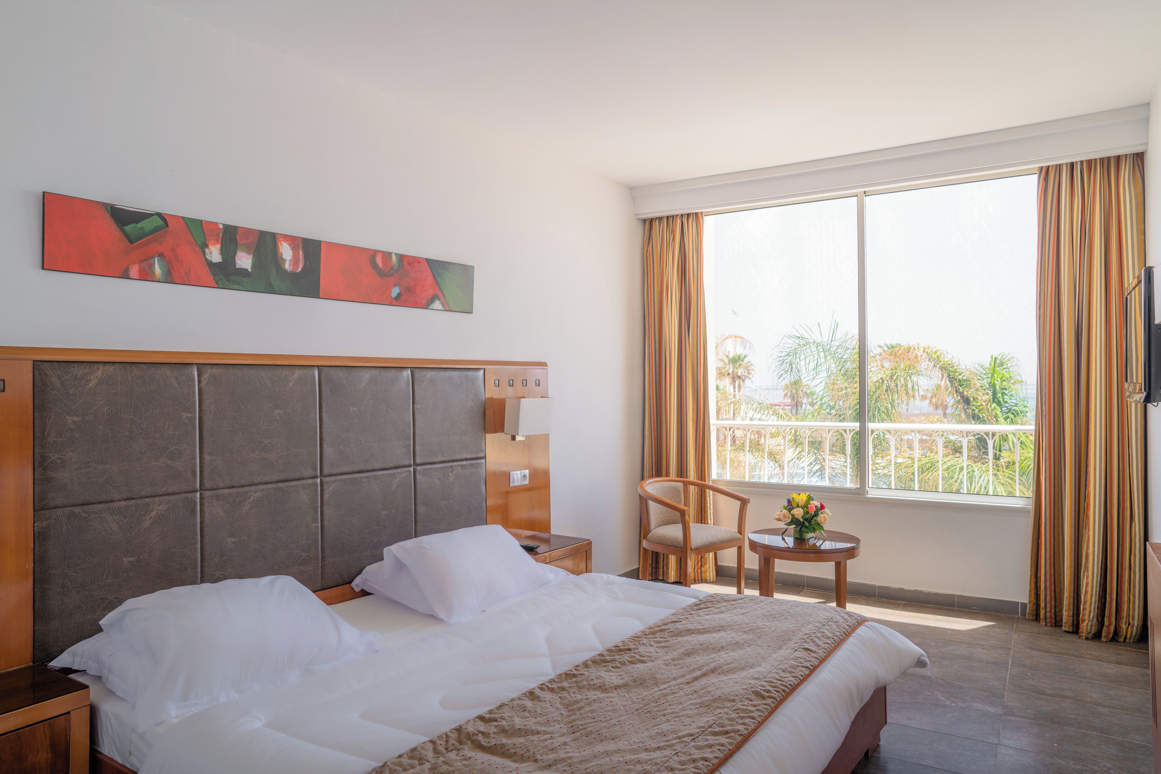 Ein Hotelzimmer mit einem großen Bett, einem Nachttisch, einem Sessel, einem kleinen Tisch mit Blumen und einem Fenster mit Vorhängen.