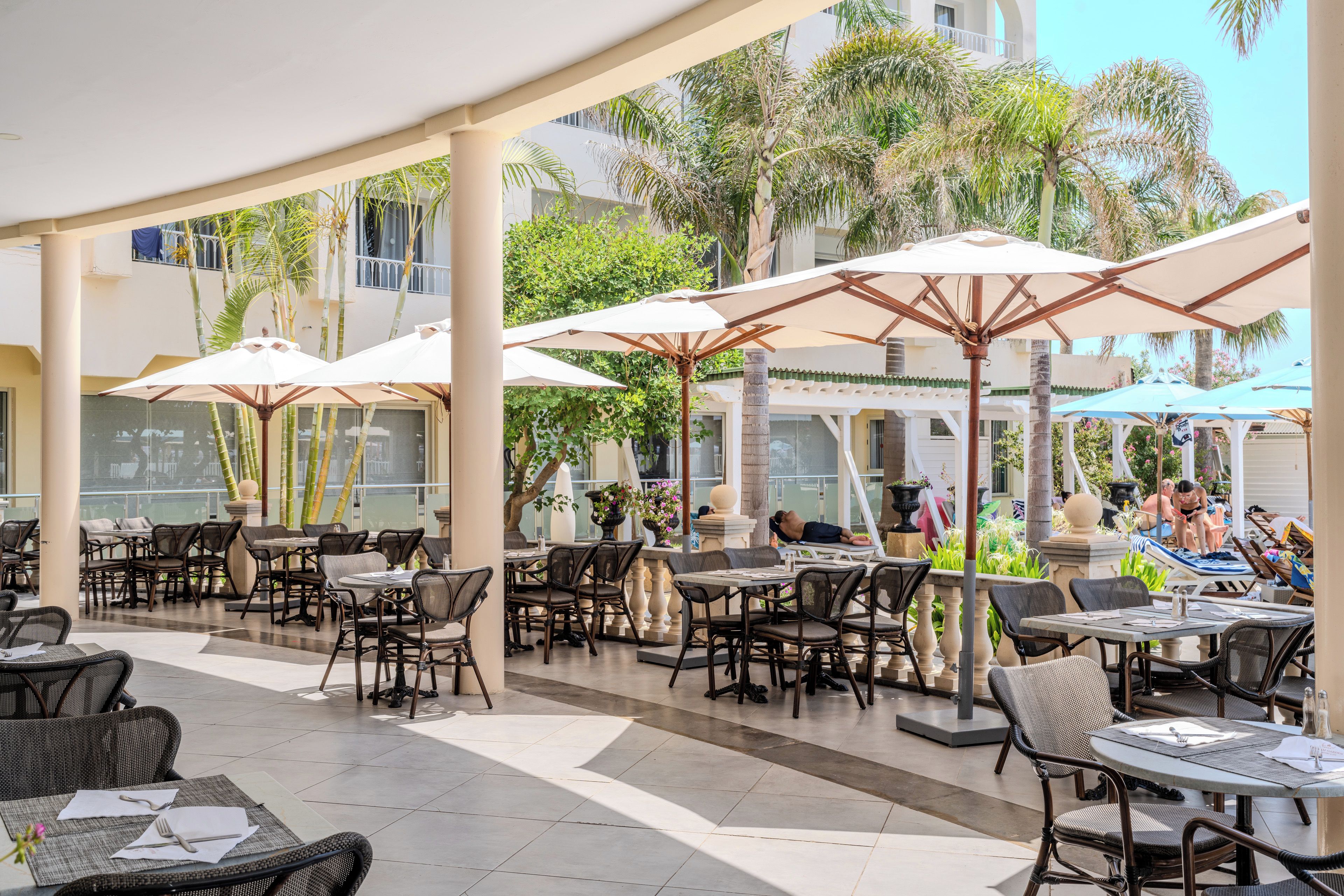 An outdoor cafe area with tables and chairs, umbrellas, and a pool area in the background.
