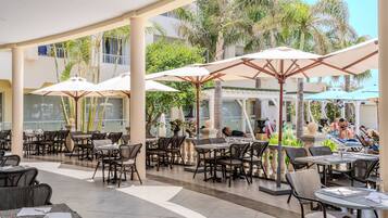 An outdoor cafe area with tables and chairs, umbrellas, and a pool area in the background.