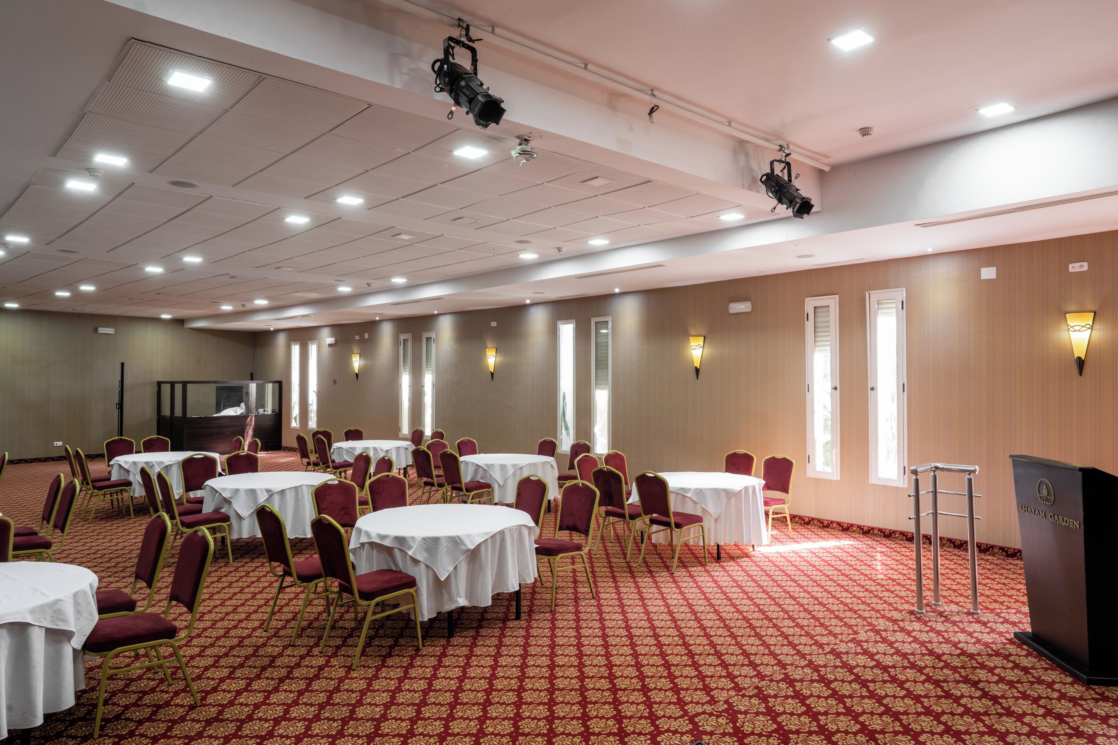 Un salón de conferencias con varias mesas y sillas, equipado con proyectores instalados en el techo.