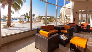 Amplio espacio interior con grandes ventanales con vistas a una zona de piscina, amueblado con mobiliario de mimbre y cojines anaranjados.
