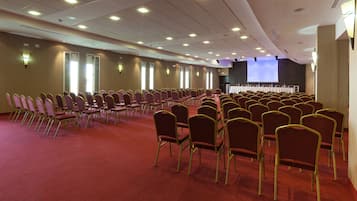 Un salón de conferencias con filas de sillas, un escenario con una pantalla de proyector y suelo alfombrado.