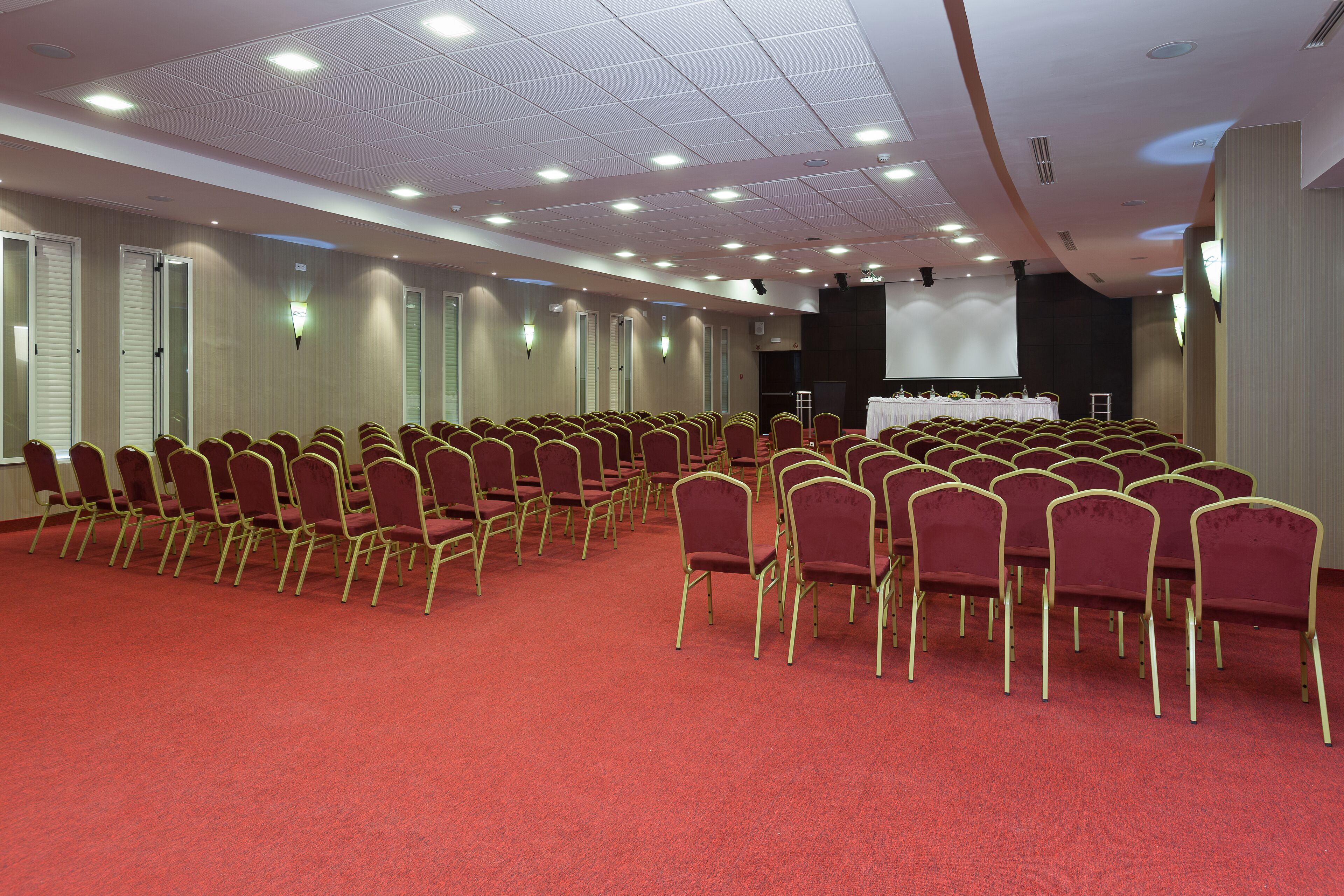 A conference room with rows of red chairs, a stage, and a projector screen.