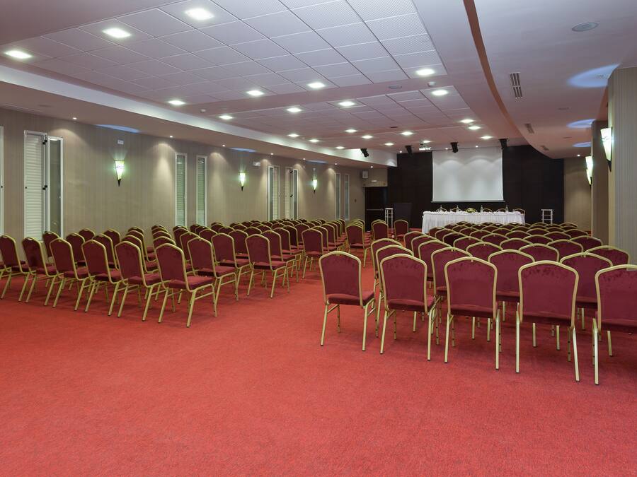 A conference room with rows of red chairs, a stage, and a projector screen.