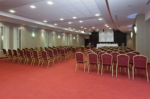Un salón de conferencias con filas de sillas rojas, un escenario y una pantalla de proyector.