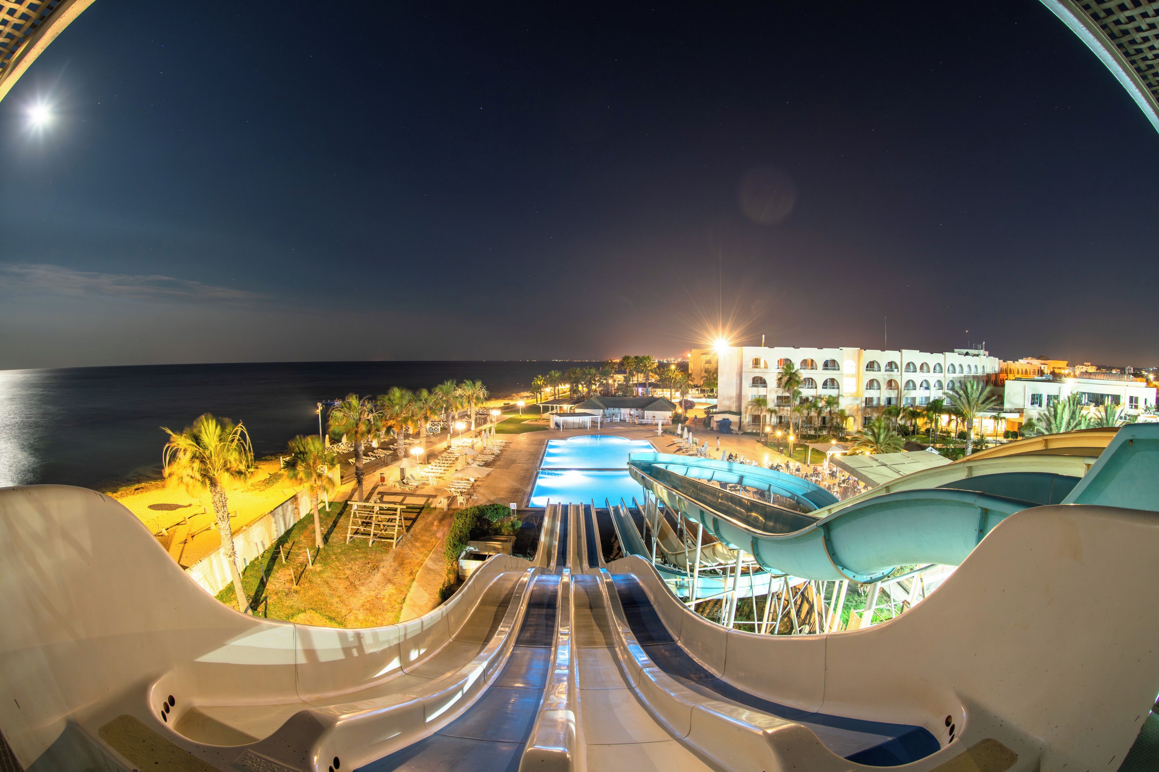 A nighttime view of a resort with a large pool, water slides, and beachfront accommodation.
