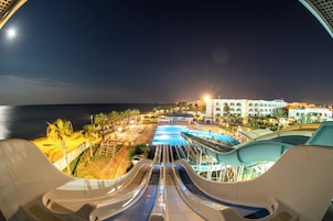 Vista nocturna de un resort con una piscina grande, toboganes acuáticos y alojamiento frente a la playa.