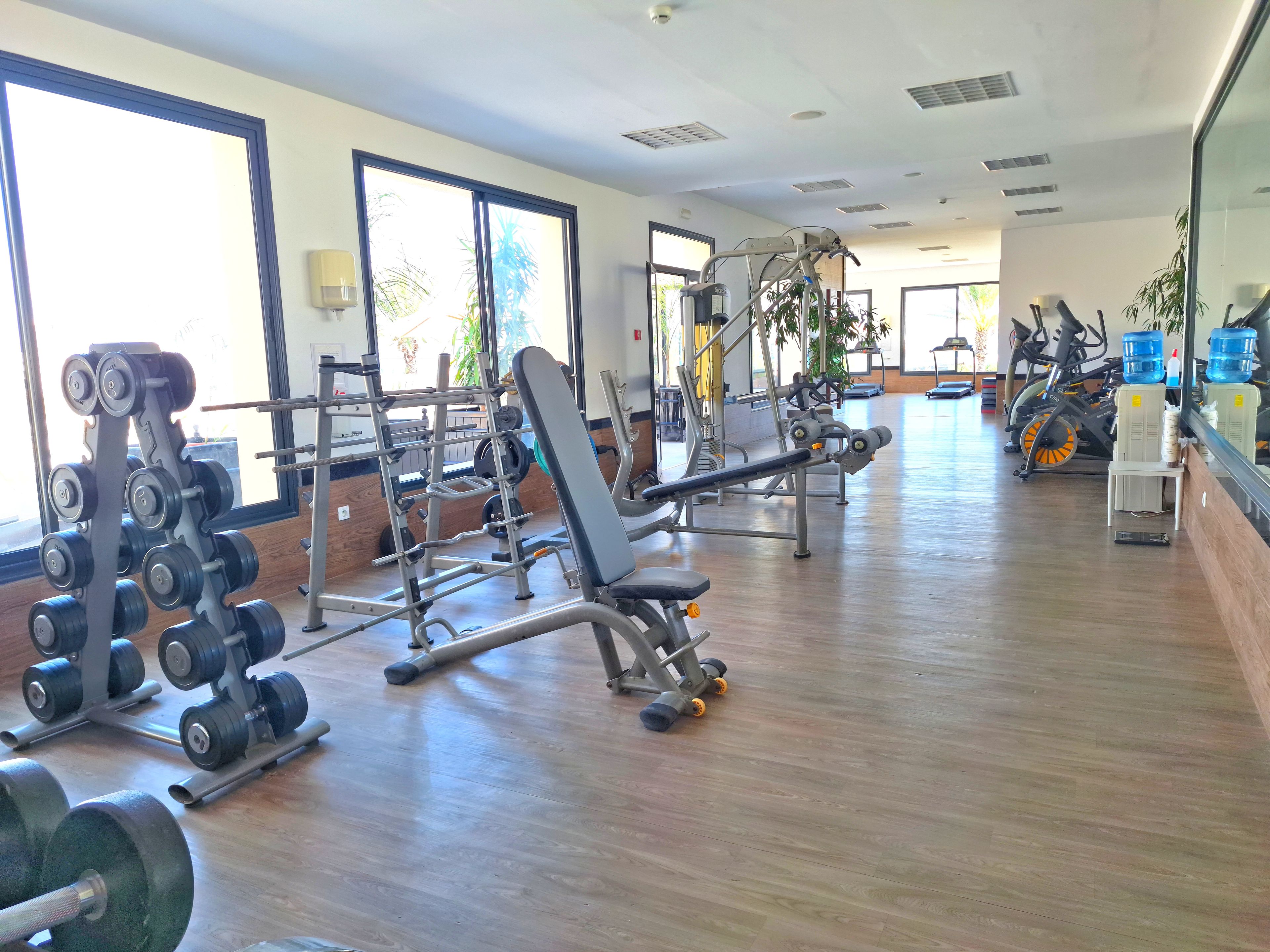 Un gimnasio con diversos equipos de ejercicio, ventanas amplias y piso de madera.