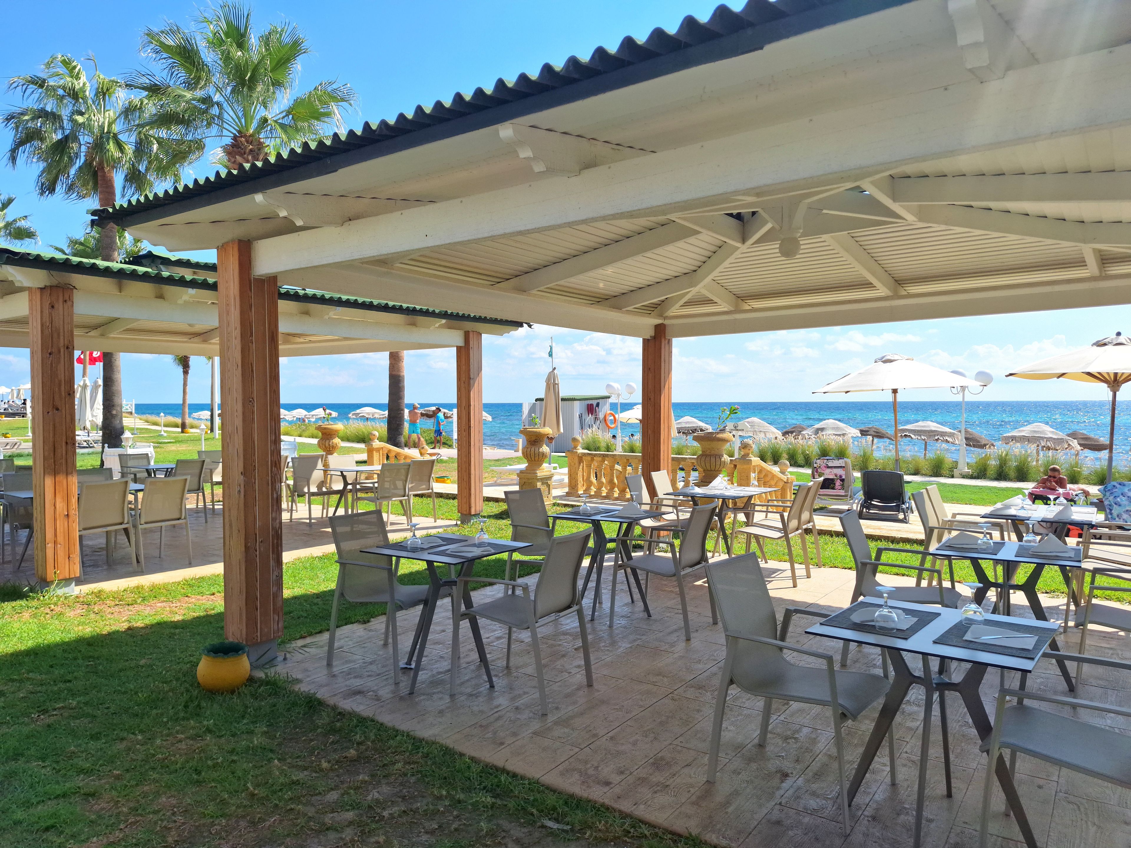 An outdoor patio with tables and chairs, overlooking a beach with umbrellas and palm trees.