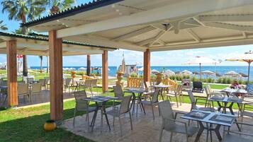An outdoor patio with tables and chairs, overlooking a beach with umbrellas and palm trees.