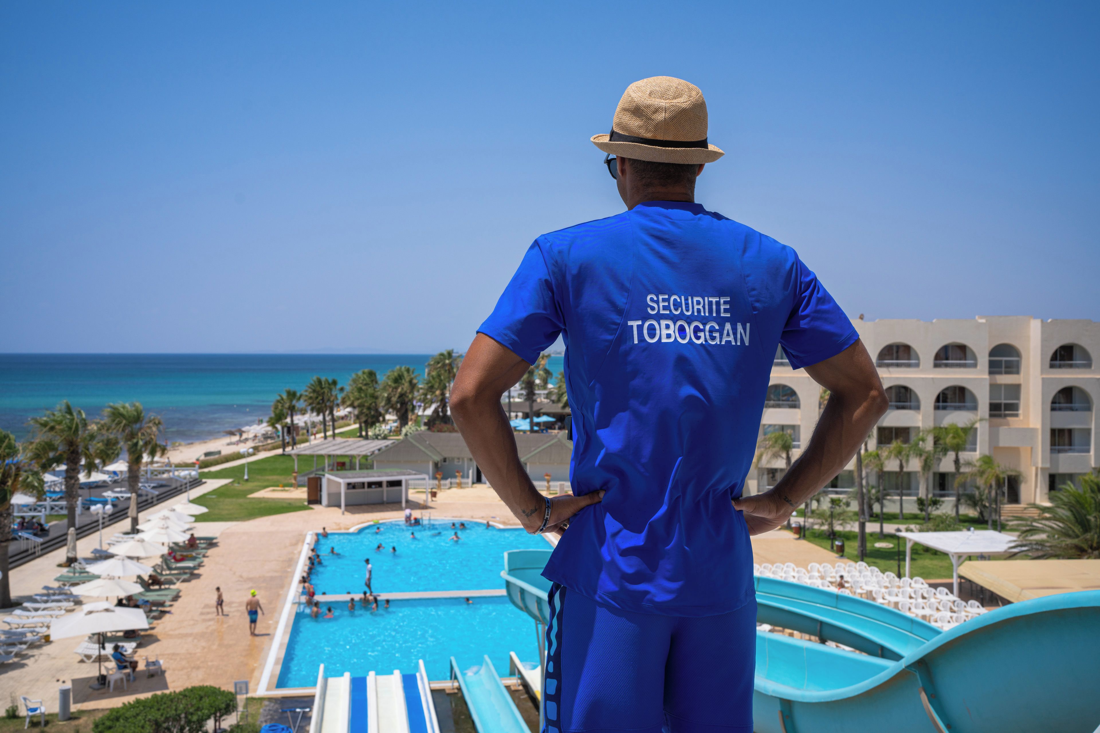Una persona con uniforme azul, con la leyenda 'SECURITE TOBOGGA' en la espalda, observa un área de piscina con vista clara a la playa y al océano.