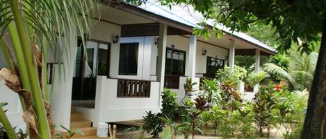 A white building with a covered porch, surrounded by lush greenery and plants.
