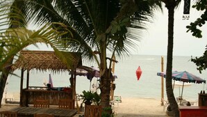 A beachside restaurant with wooden tables and chairs, a thatched roof structure, and umbrellas. - Marina Beach Resort (Koh Samui)