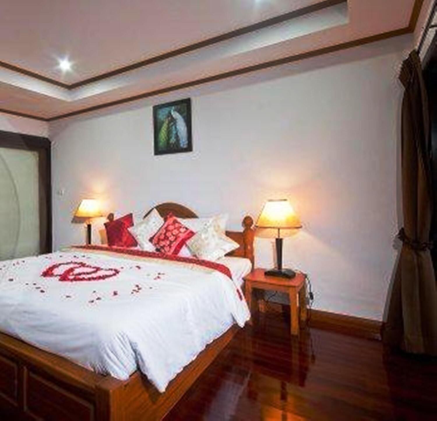 A neatly made bed with a white bedspread and red heart-shaped decoration, two bedside lamps, a wooden headboard, and a painting on the wall.