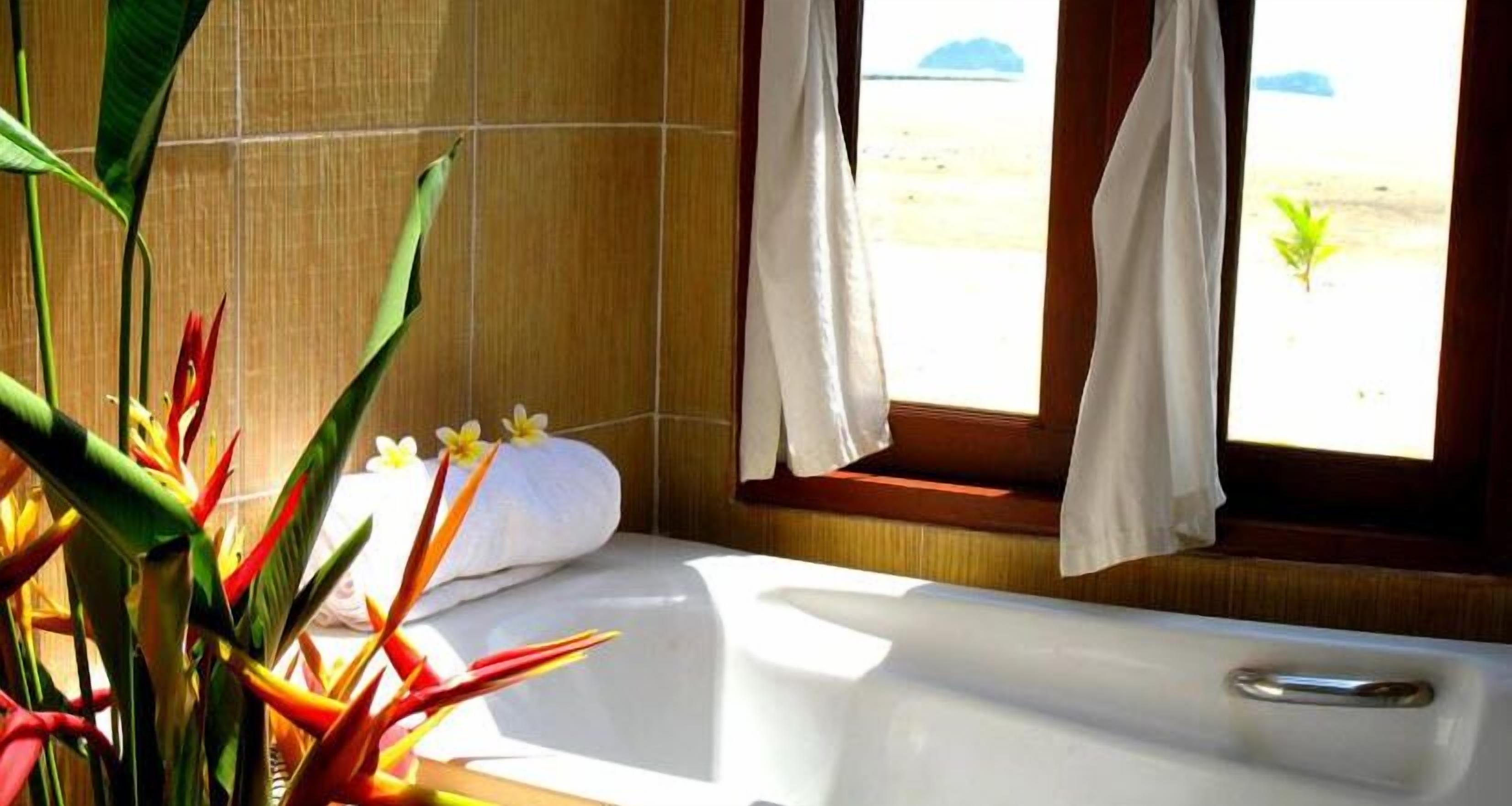 A bathroom with a bathtub, a window with curtains, and a plant with red and yellow flowers.