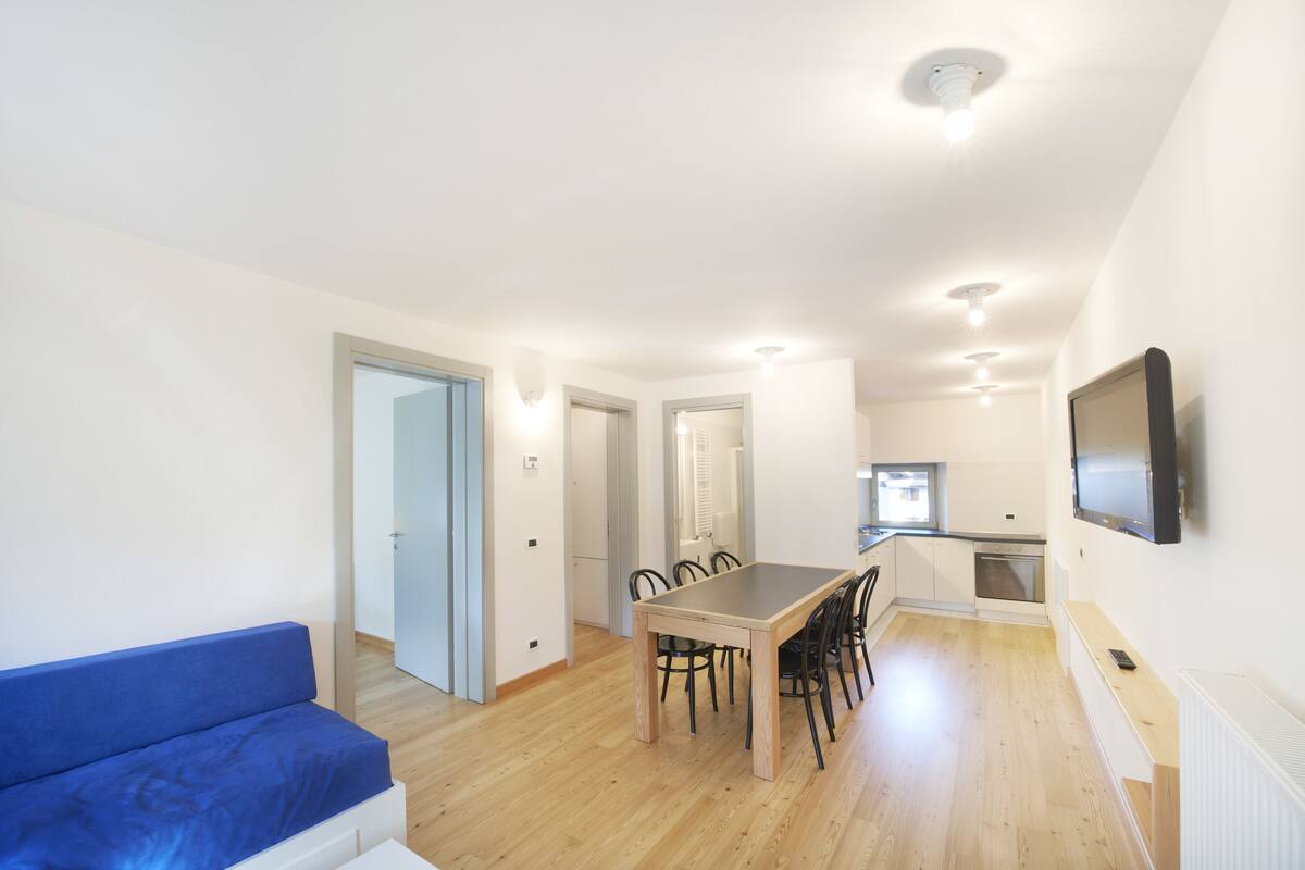 A modern apartment with a dining area, a sofa, a TV mounted on the wall, and a kitchen in the background.