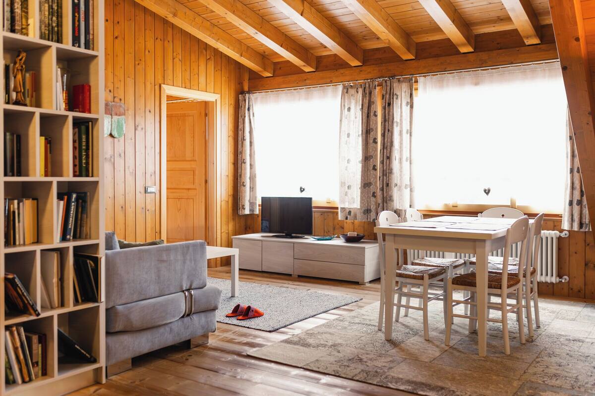 A cozy living room with a wooden interior, a bookshelf, a grey sofa, a dining table with chairs, and a television.