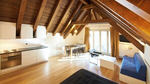 Un loft moderno con vigas de madera a la vista, una cocina con armarios blancos, una zona de comedor con mesa y sillas, y una sala de estar con un sofá azul y una alfombra negra.