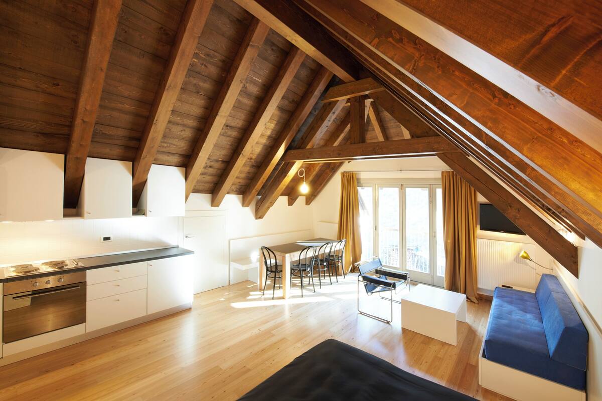 A modern loft apartment with exposed wooden beams, a kitchen with white cabinetry, a dining area with a table and chairs, and a living space with a blue sofa and a black rug.