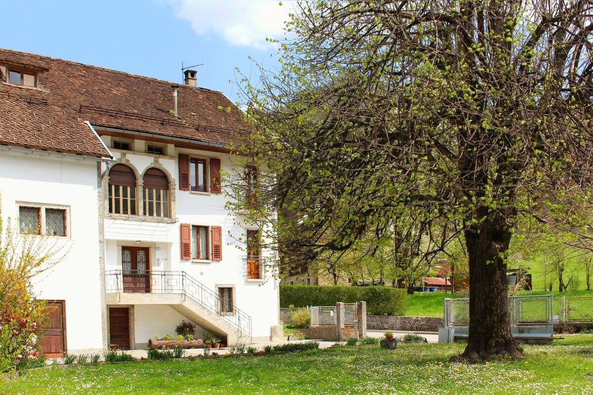 A two-story white building with a tiled roof, a balcony, and a tree in the foreground.