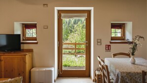 Una habitación con puerta de madera que da a un balcón, un televisor sobre un mueble de madera, una mesa de comedor con sillas y una ventana pequeña con cortina.
