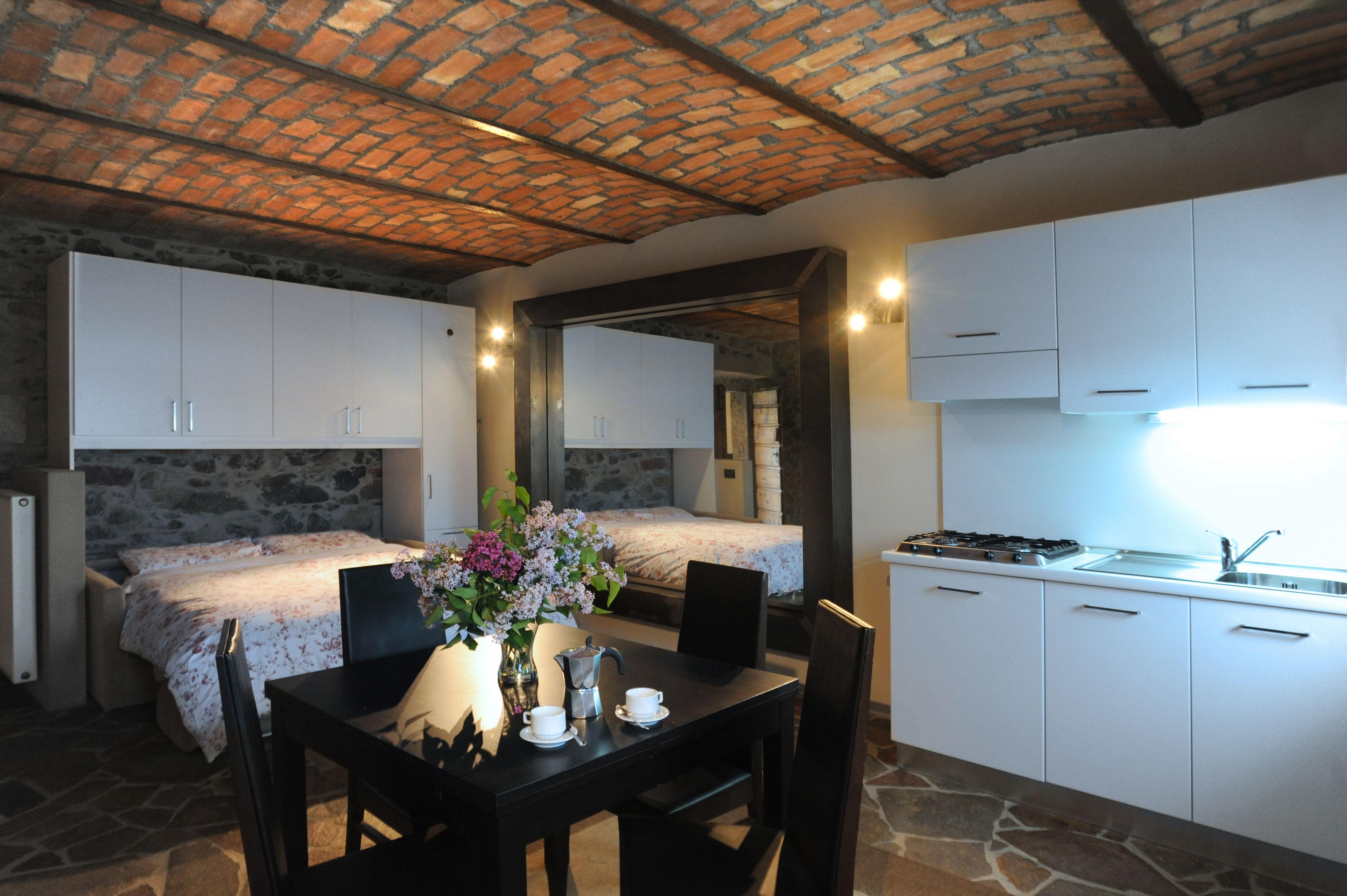 Habitación pequeña con cama, una mesa de comedor con sillas, una zona de cocina con armarios y una pared de piedra.