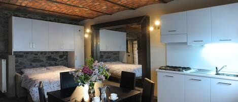 Habitación pequeña con cama, una mesa de comedor con sillas, una zona de cocina con armarios y una pared de piedra.