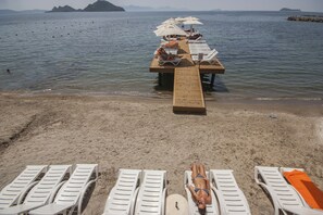 A beach with sun loungers and umbrellas, a wooden pier extending into the water, and a clear view of the sea and distant islands.