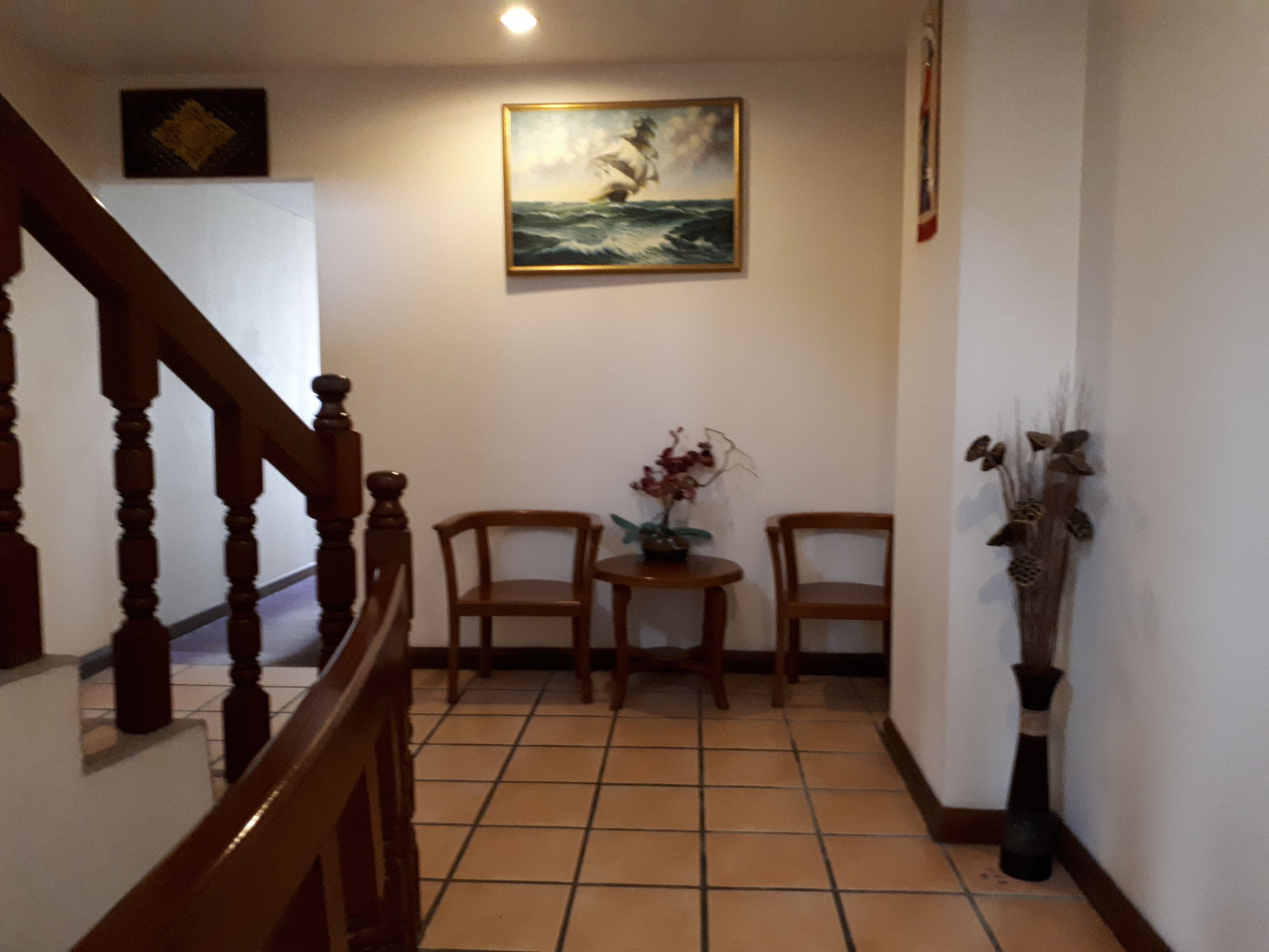 Un couloir doté d’un sol carrelé, d’un escalier en bois, de deux chaises en bois, d’une petite table sur laquelle se trouve un vase, et d’un tableau représentant un voilier accroché au mur.