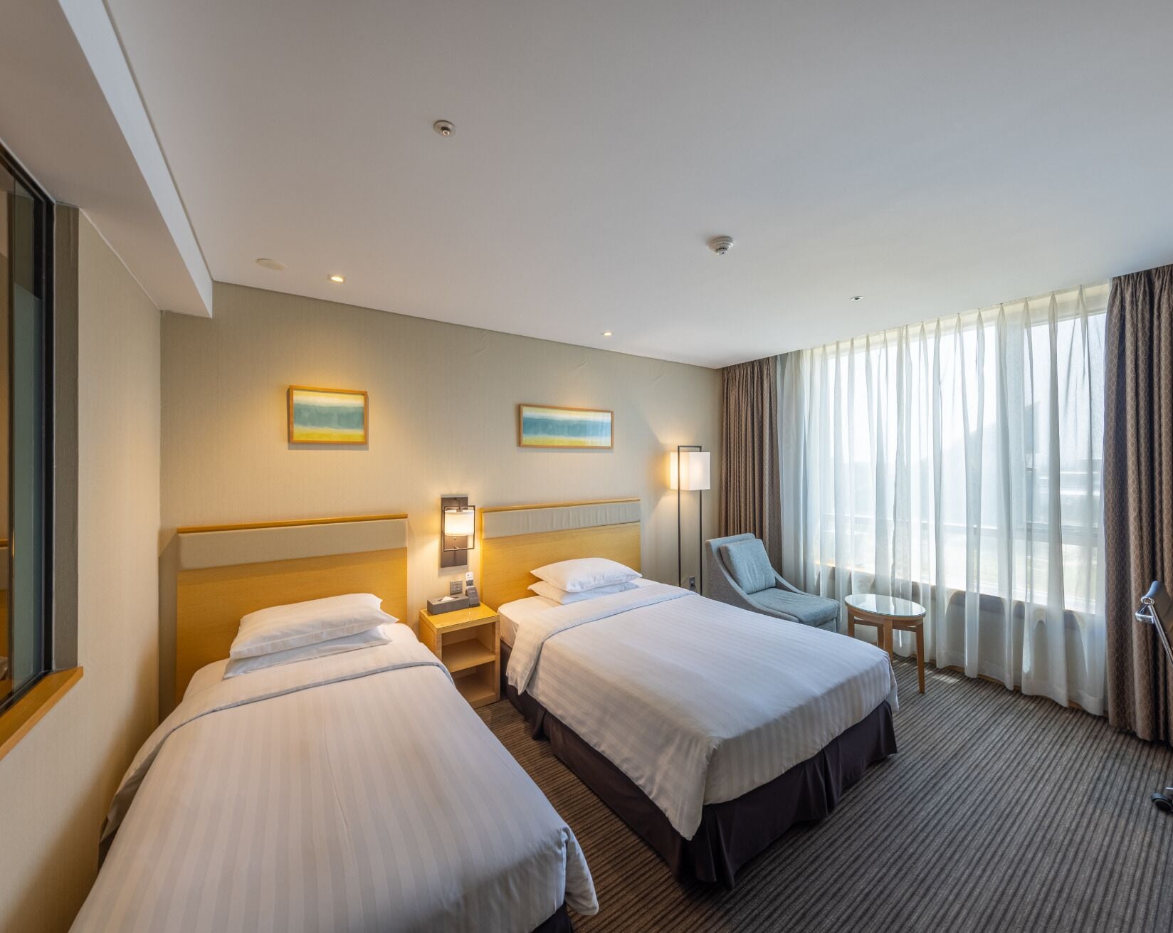 Quarto de hotel com duas camas, uma escrivaninha, uma cadeira, um abajur e uma janela com cortinas.
