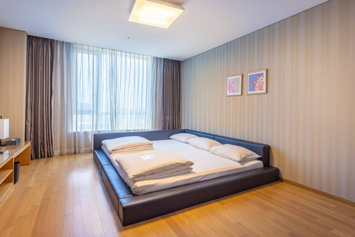 A modern hotel room with a large bed, wooden flooring, a desk with a lamp, and a large window with curtains.