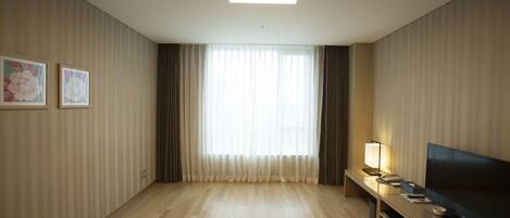 Una habitación de hotel moderna con piso de madera, un televisor de pantalla plana grande, una mesita de noche con lámpara y una ventana con cortinas traslúcidas.