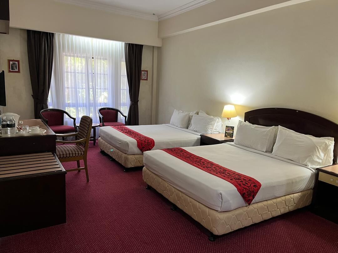 A hotel room with two beds, a desk, chairs, a lamp, and a window with curtains.