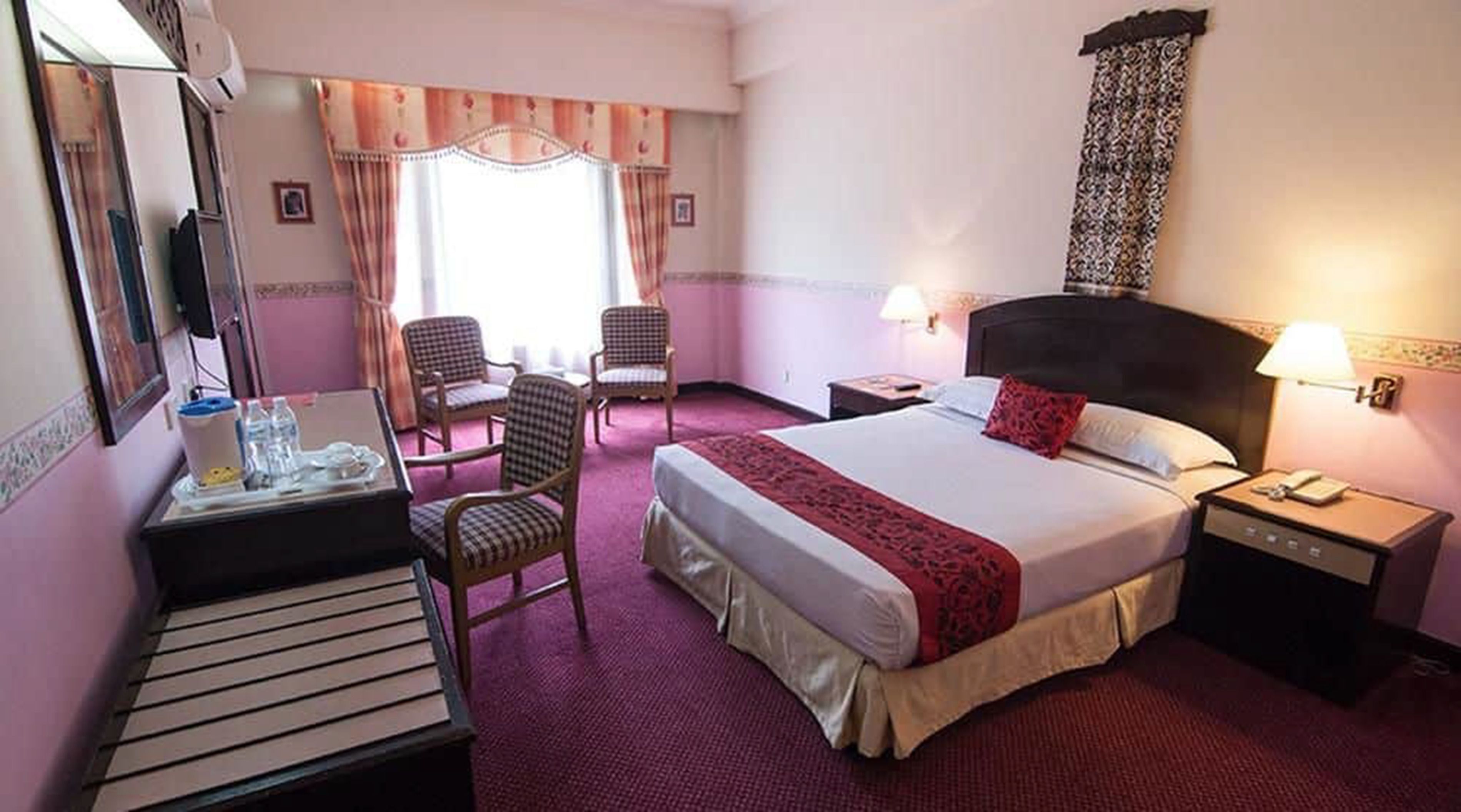 Une chambre d’hôtel avec un lit, deux chaises, un bureau, une table de chevet et une fenêtre avec des rideaux.