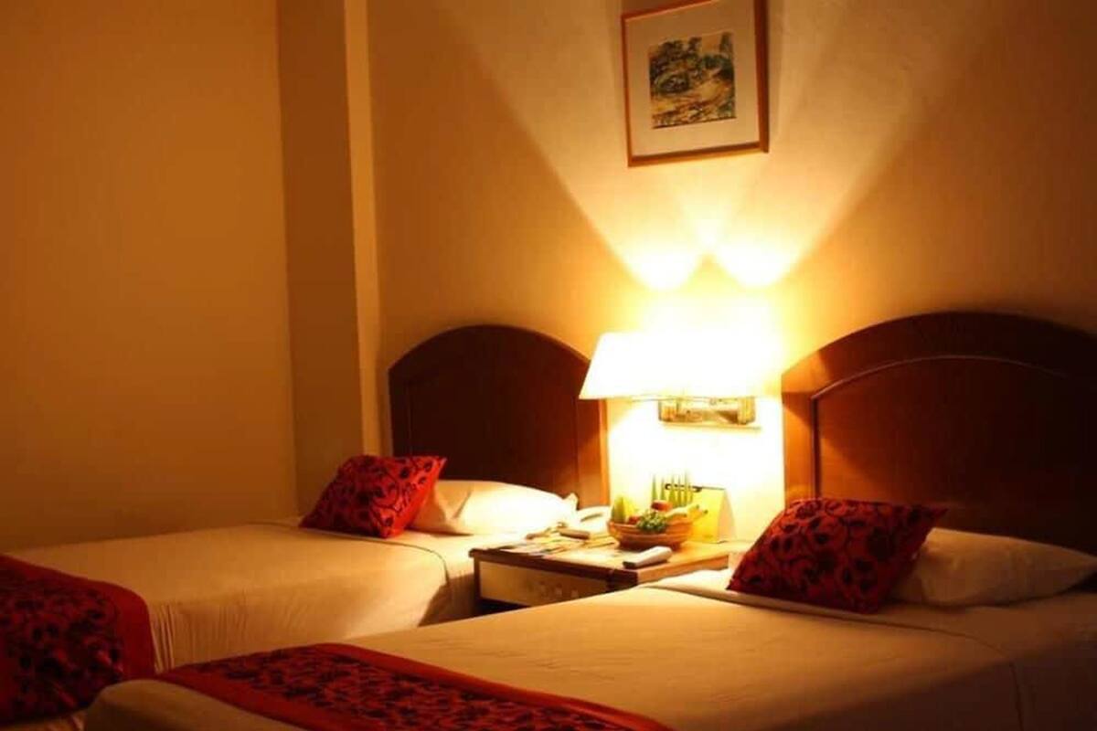 A hotel room with two beds, each with a red patterned throw blanket, a bedside table with a lamp, and a framed picture on the wall.