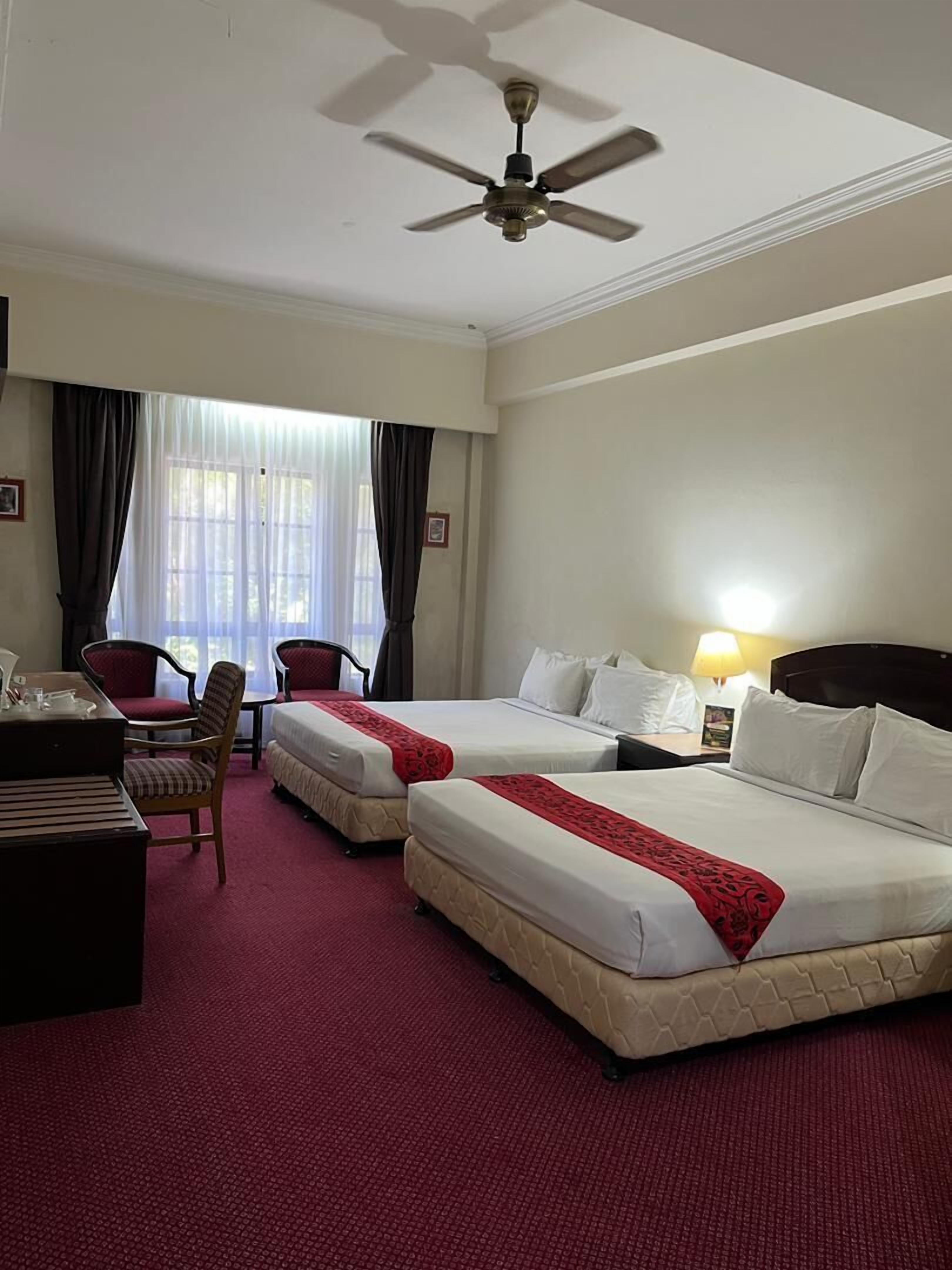 A hotel room with two beds, a desk, a chair, a piano, and a ceiling fan.