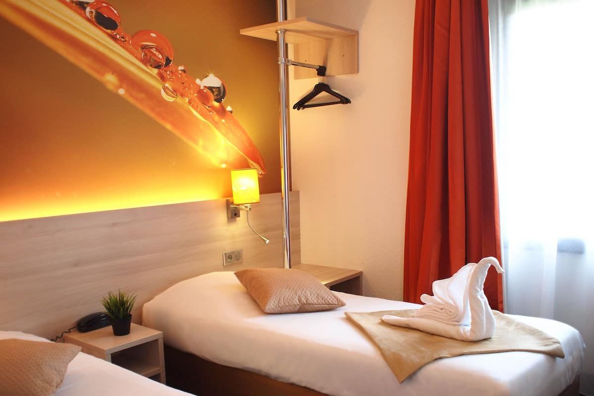 Ein Hotelzimmer mit zwei Betten, einem Nachttisch mit Lampe, einer Wandleuchte und einem Fenster mit Vorhängen.