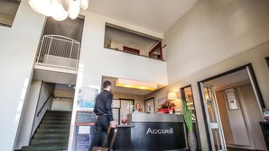 Een hotel receptie met een zwart receptiebalie waarop 'Accueil' staat, een trap en een grote kroonluchter.