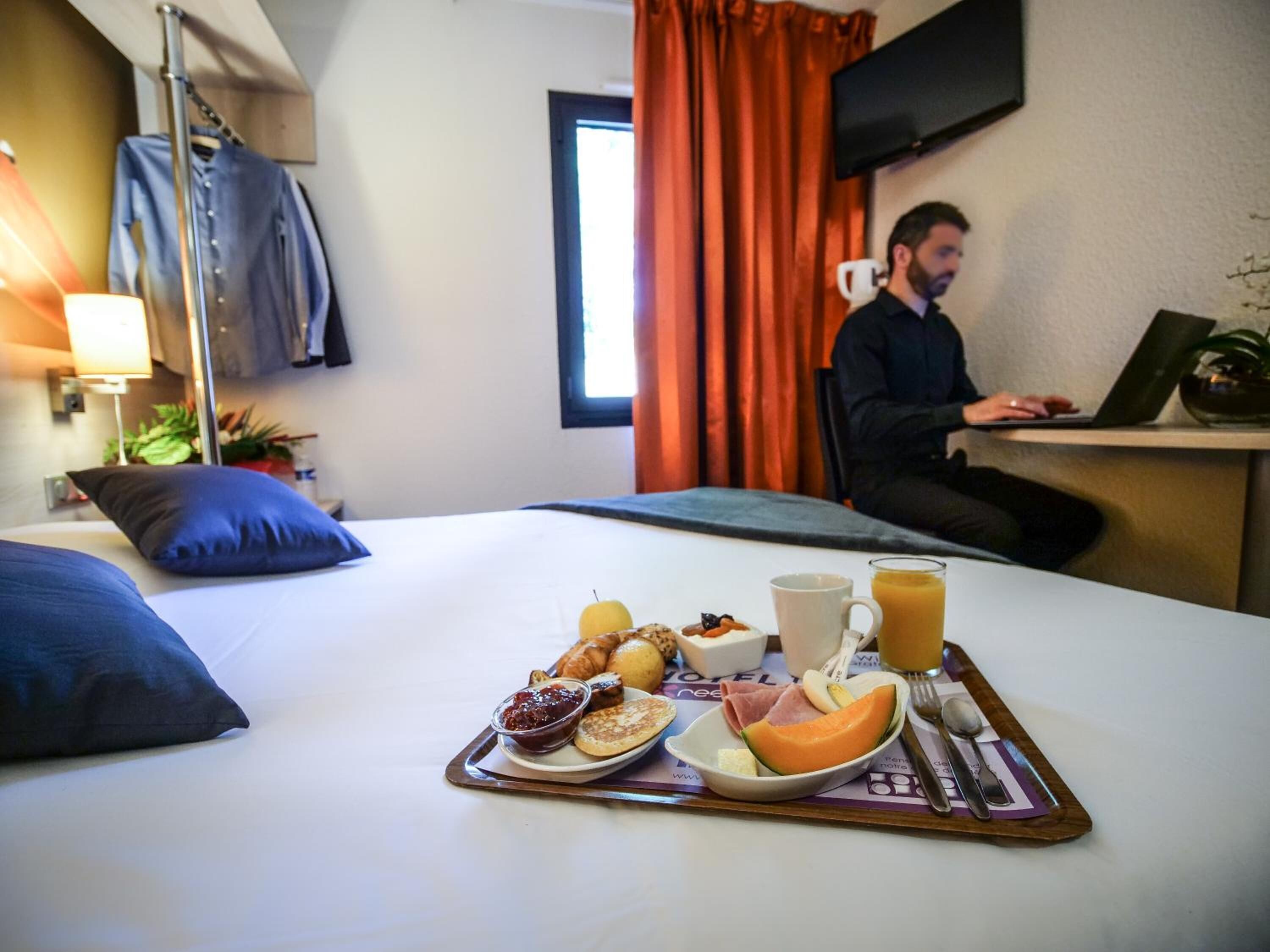 A hotel room with a bed, breakfast tray, and a person working at a desk.