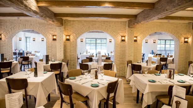 Restaurante com paredes de tijolo, mesas brancas e cadeiras marrons.
