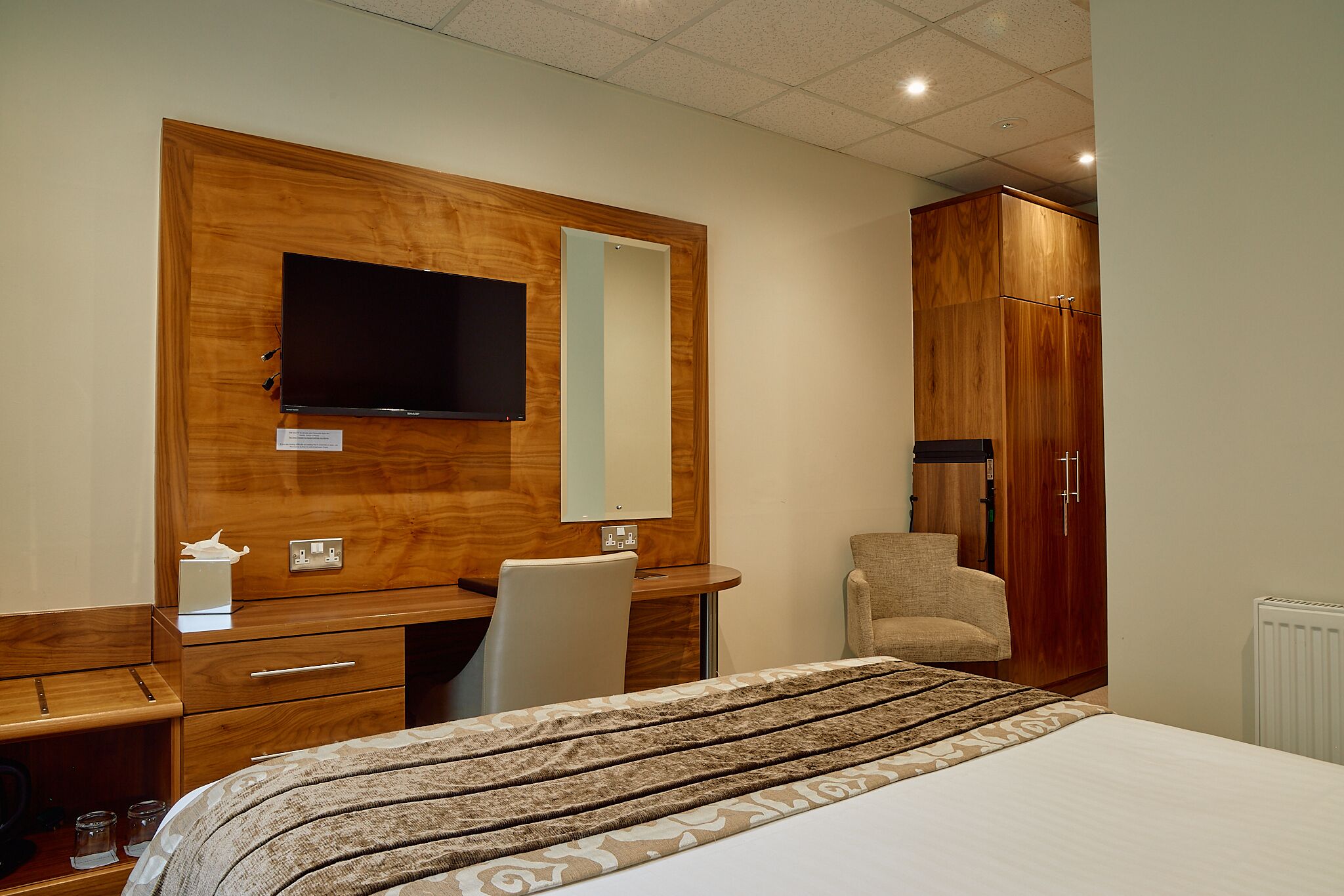 A hotel room with a wooden desk, a television, a chair, and a wardrobe.