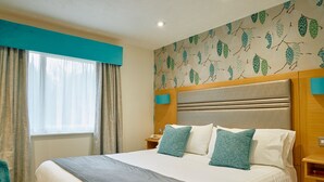 Quarto de hotel com uma cama grande, mesa de cabeceira, janela com cortinas e parede com padrão de folhagem.