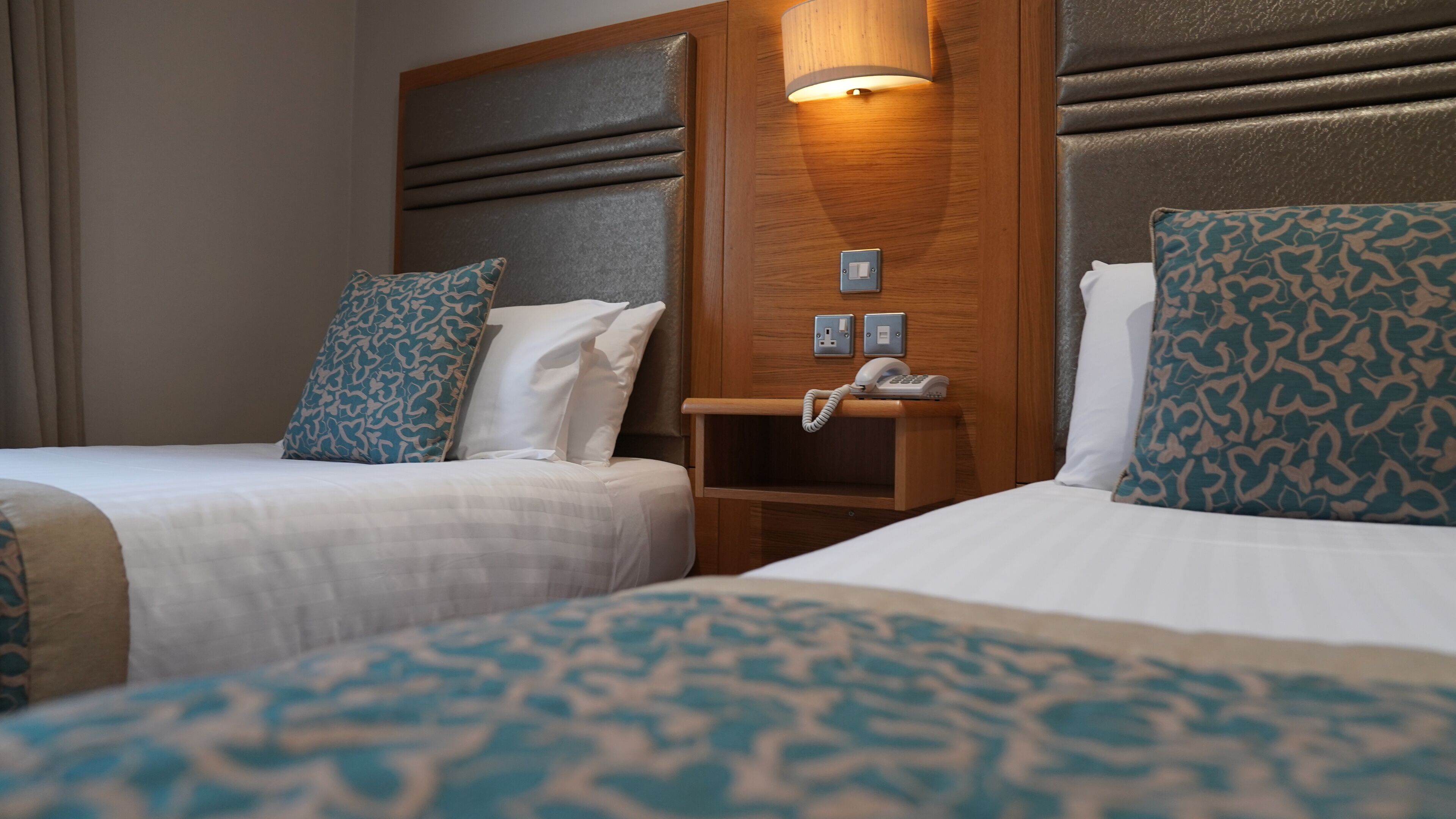 A hotel room with two beds, each with patterned bedding and pillows. There is a wooden headboard with a built-in phone and light fixture.